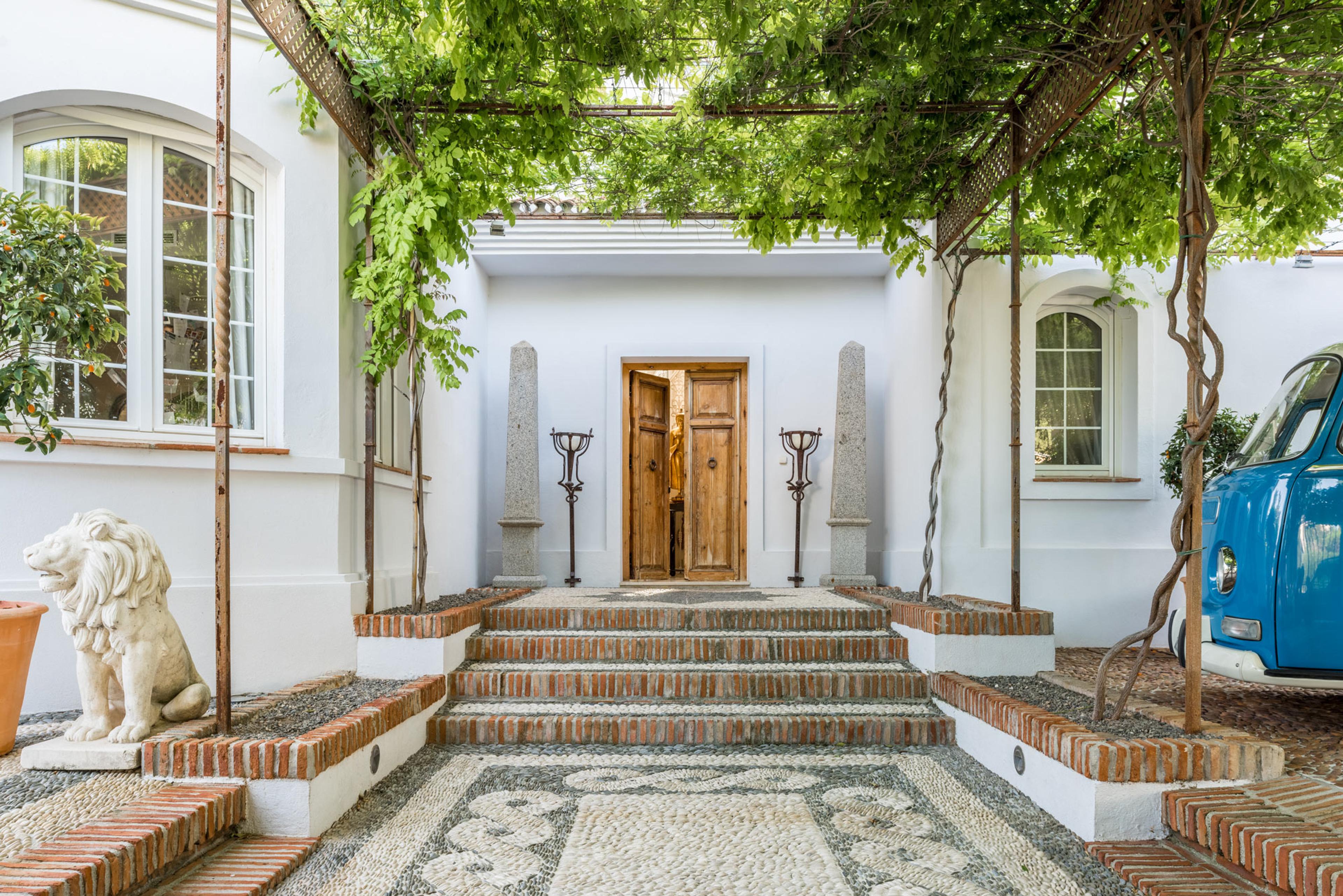 Charming Mediterranean entrance with rustic wooden door, brick steps, pebble mosaic, greenery, stone lion statue, and vintage blue vehicle.