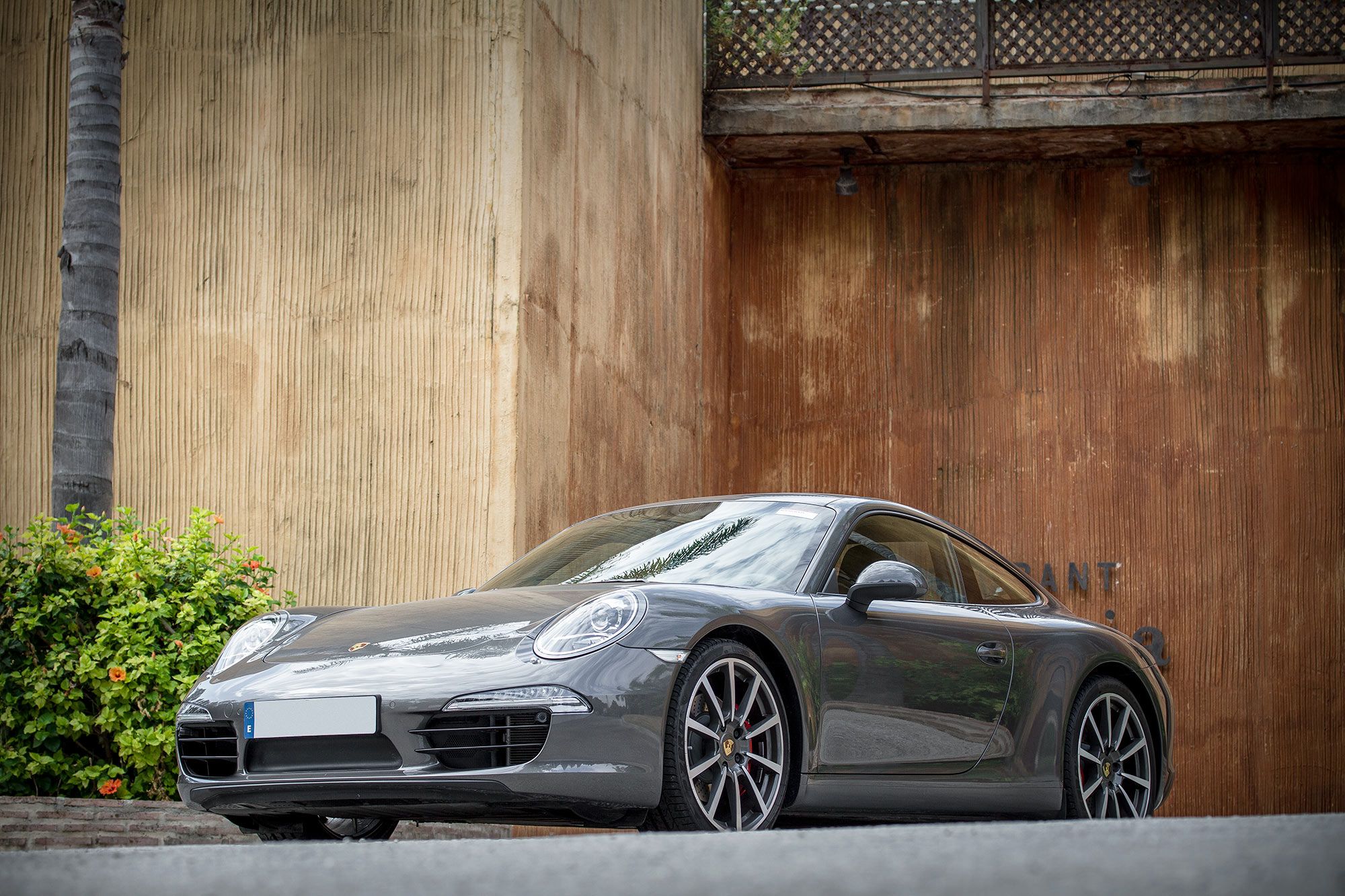 Luxury gray sports car parked against a rustic textured wall, surrounded by tropical greenery and a modern minimalist backdrop.
