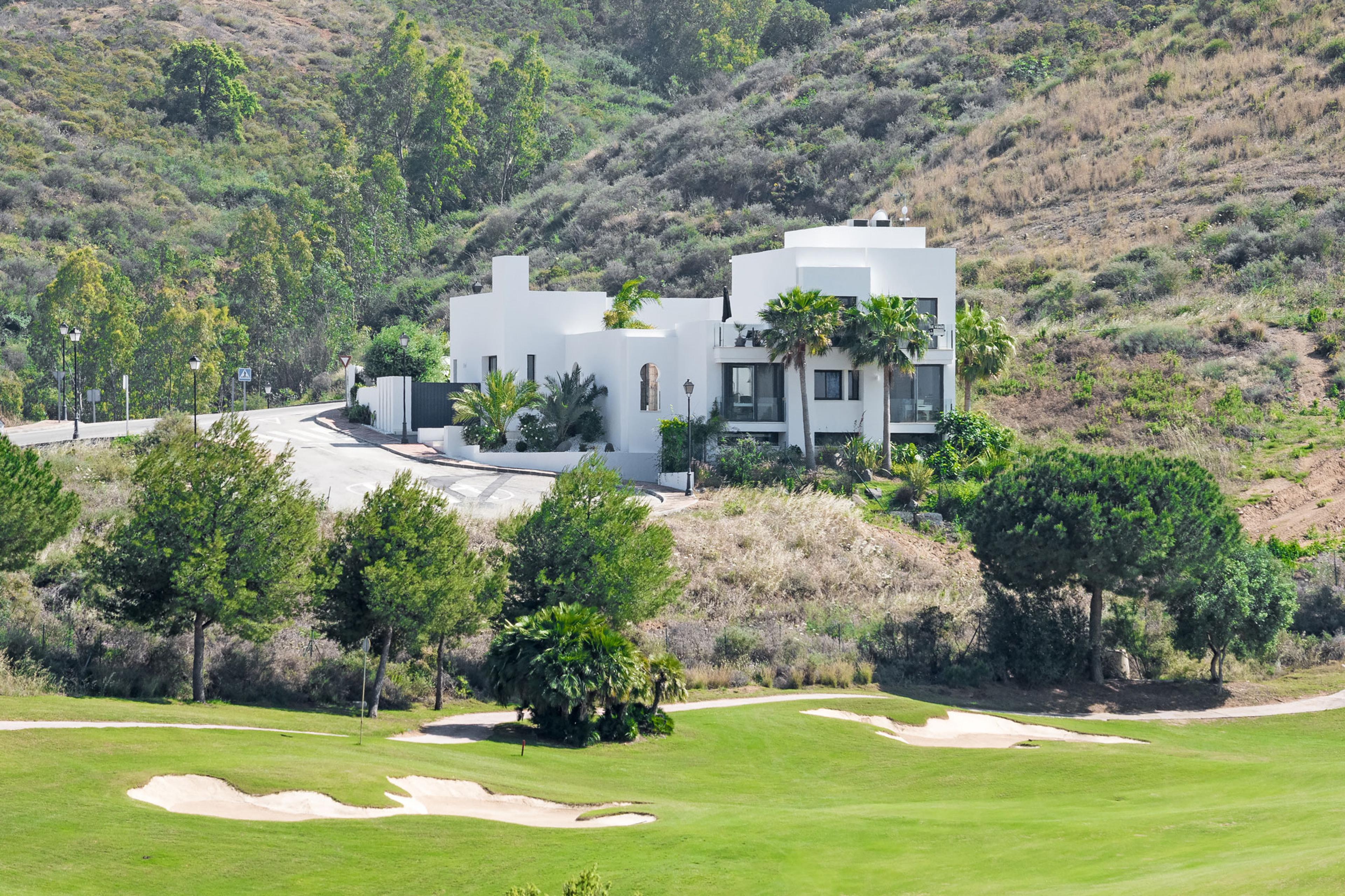 Modern white villa surrounded by lush greenery and a golf course, set against a backdrop of rolling hills.