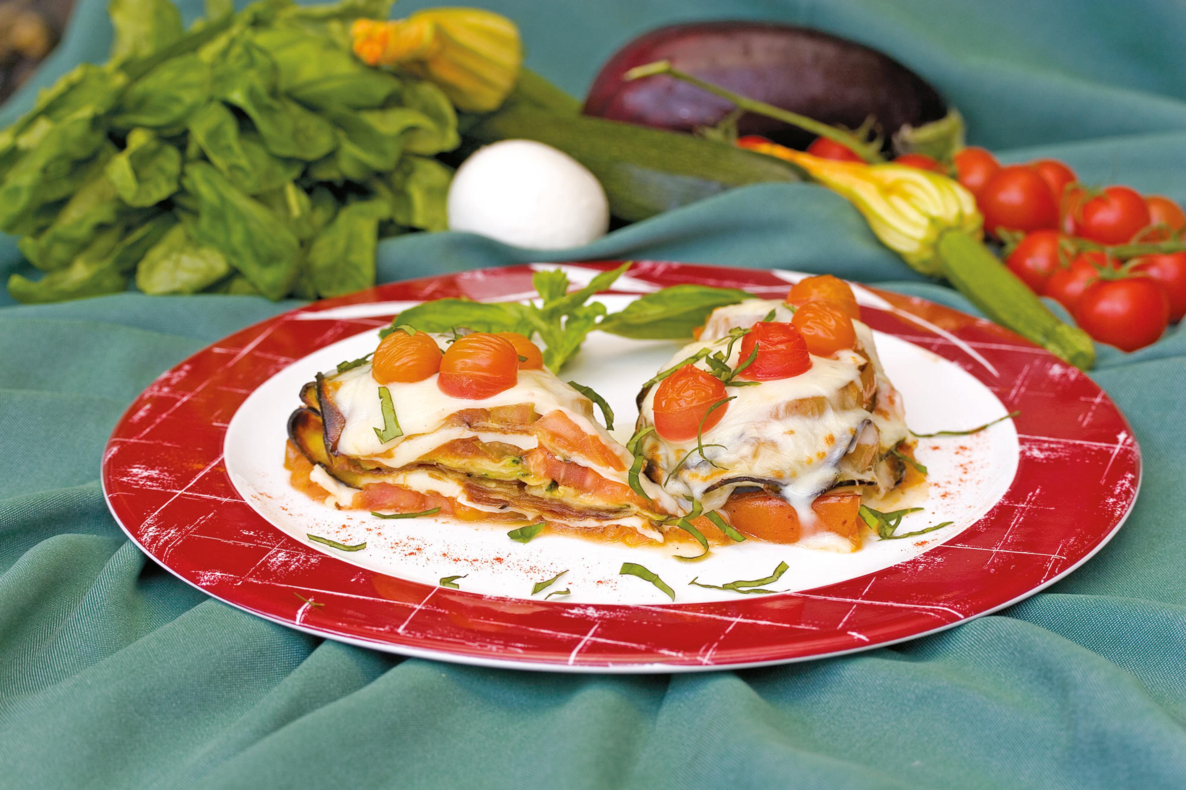 Vegetable lasagne topped with melted cheese and cherry tomatoes, served on a red plate with fresh vegetables in the background.