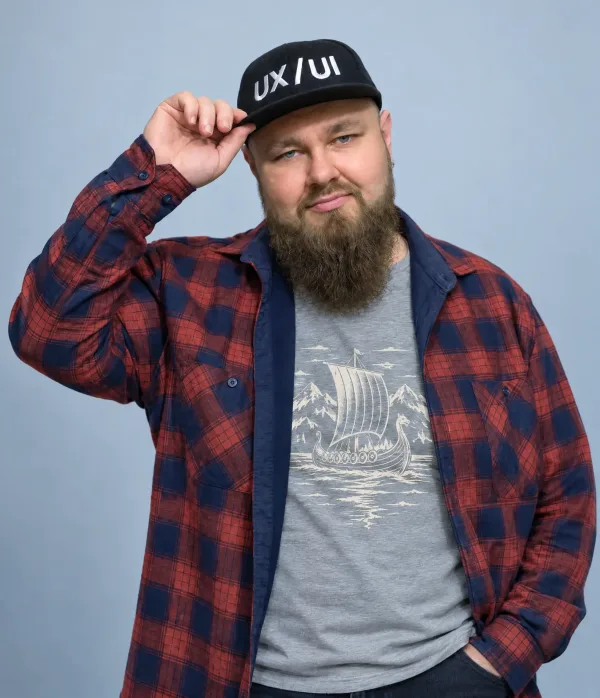 Serhii Lanchukovskyi tipping the brim of a cap that reads UX/UI, plaid shirt over a tee with a Viking ship, plain light-blue background