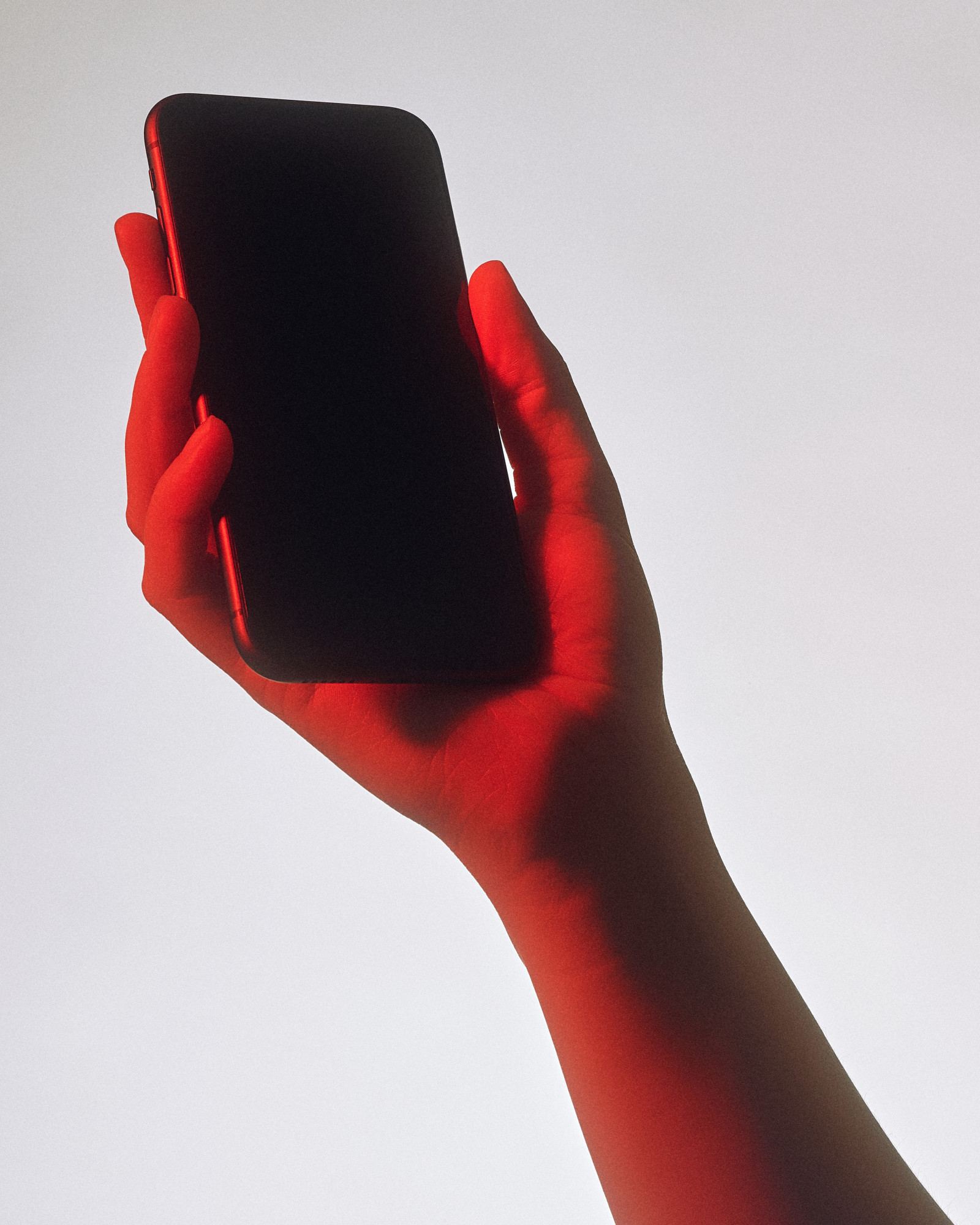 Uma mão iluminada de vermelho, erguida sobre um fundo cinza claro, segurando um celular de tela preta e apagada