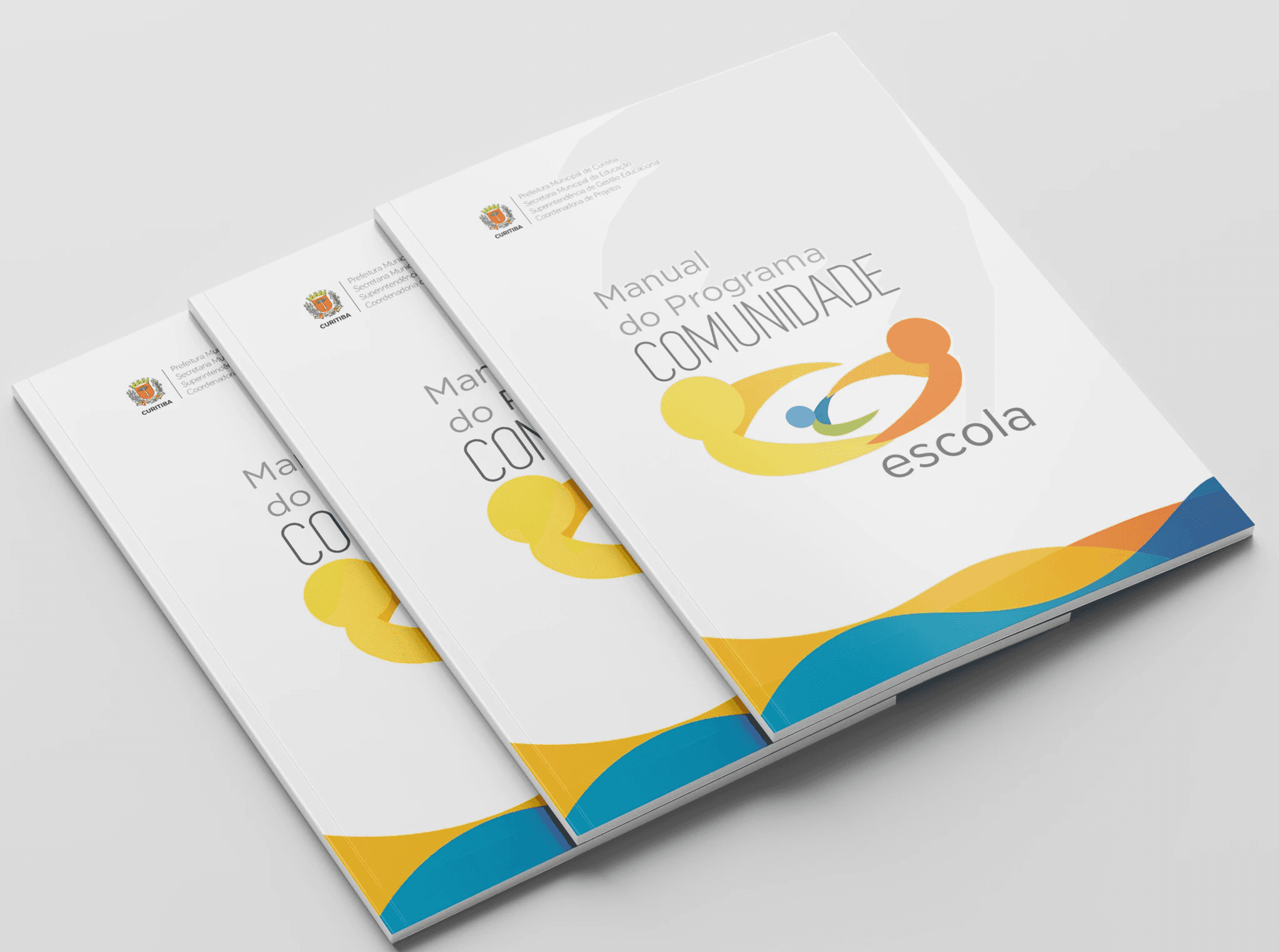 Três exemplares do Manual do Programa Comunidade Escola em leque, com o logo em forma de olho amarelo, laranja, azul e verde