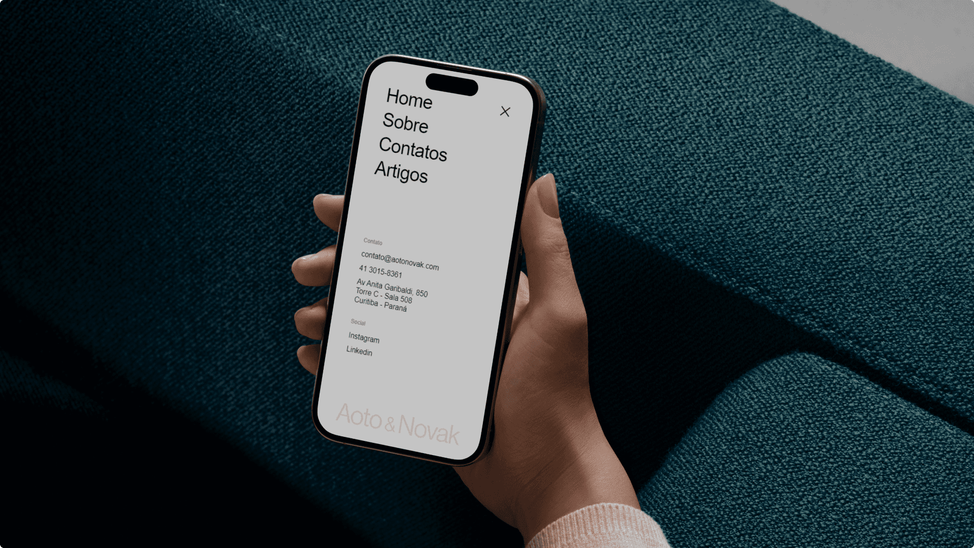 Mão segura o celular sobre sofá verde-azulado com o menu da Aoto & Novak aberto: Home, Sobre, Contatos, Artigos