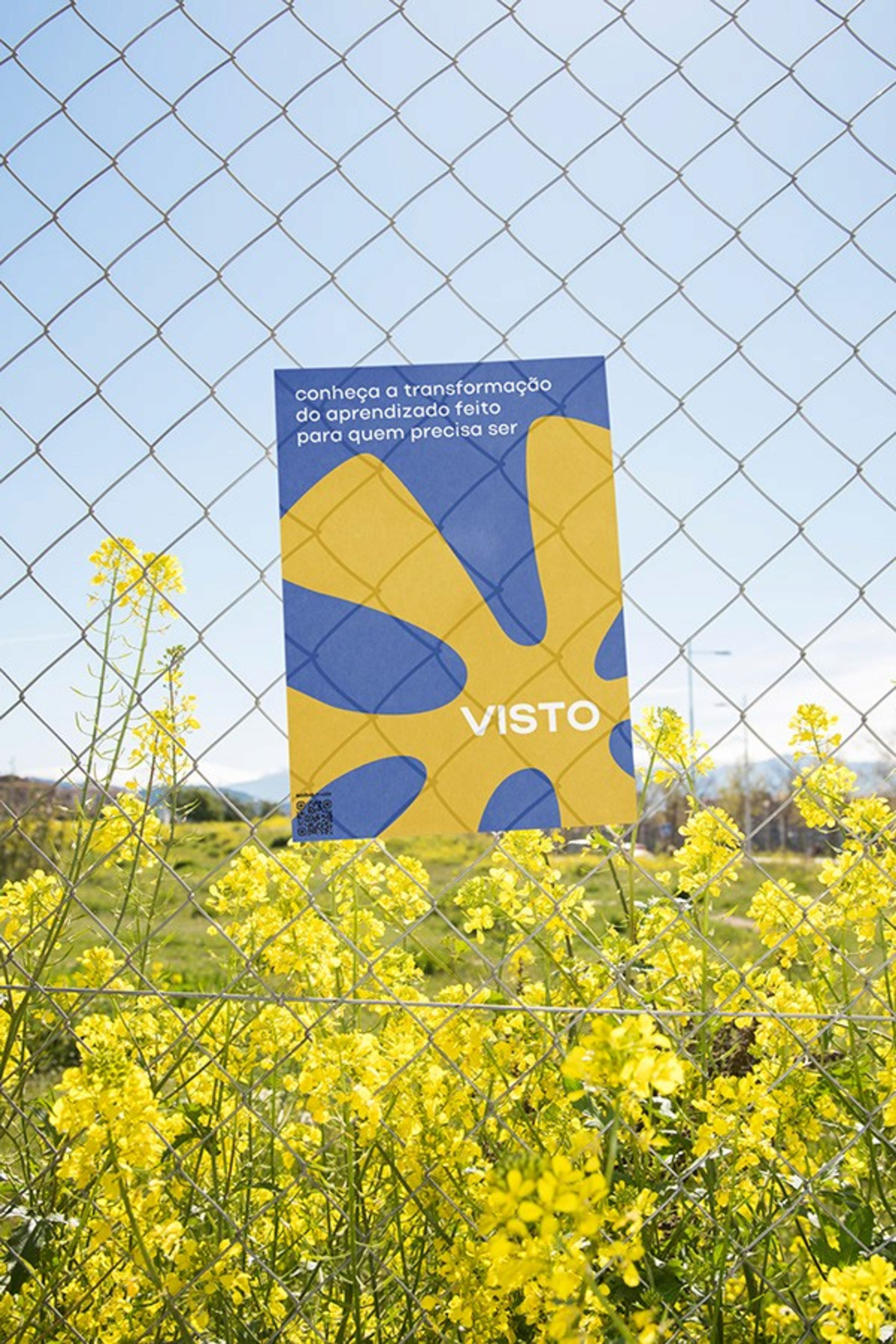 Cartaz azul e amarelo preso ao alambrado sobre flores amarelas: 'para quem precisa ser VISTO'
