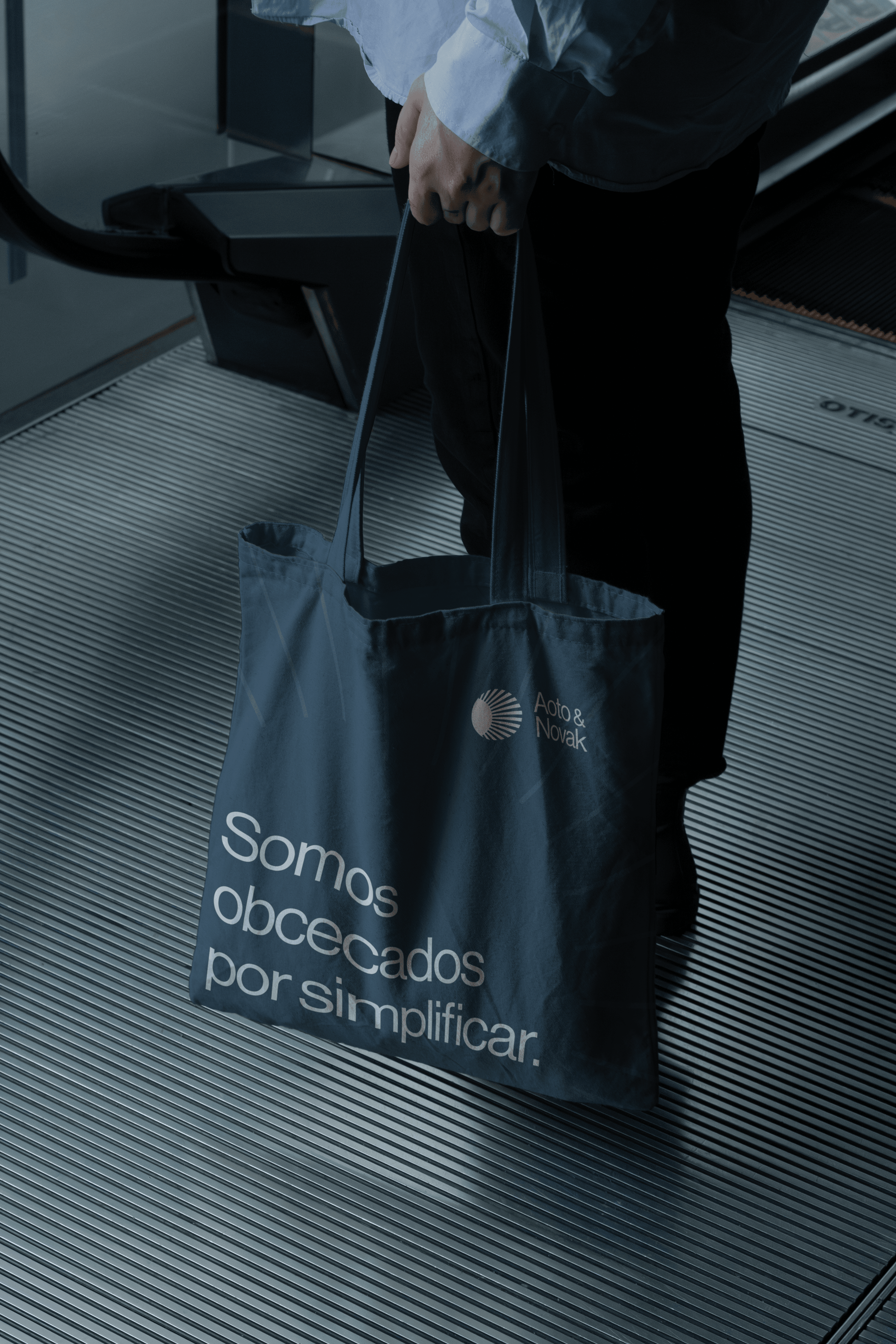 Ecobag azul-petróleo da Aoto & Novak com Somos obcecados por simplificar, carregada no pé da escada rolante