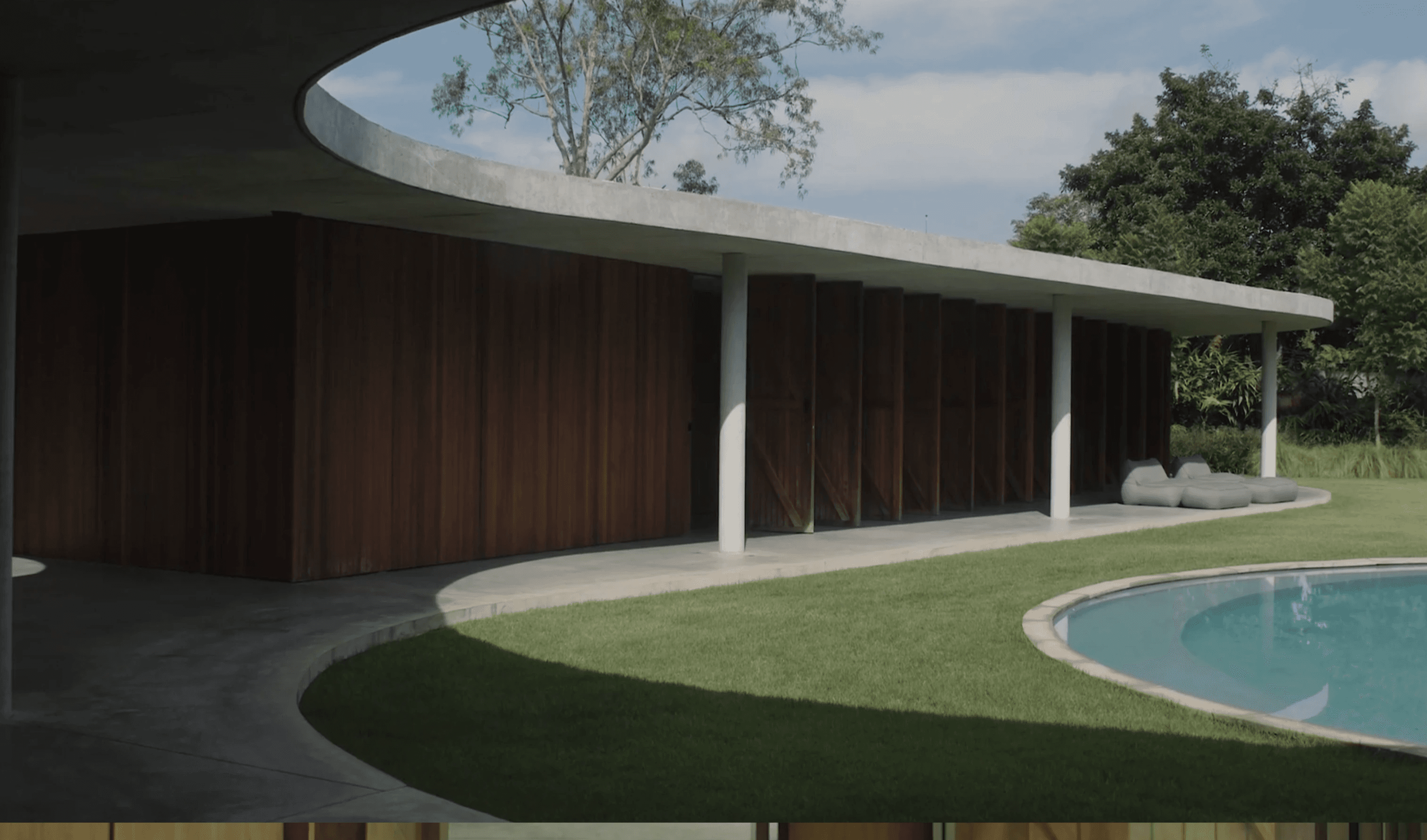 Marquise de concreto curva sobre fachada de madeira escura, colunas brancas finas e futons cinza voltados para a piscina