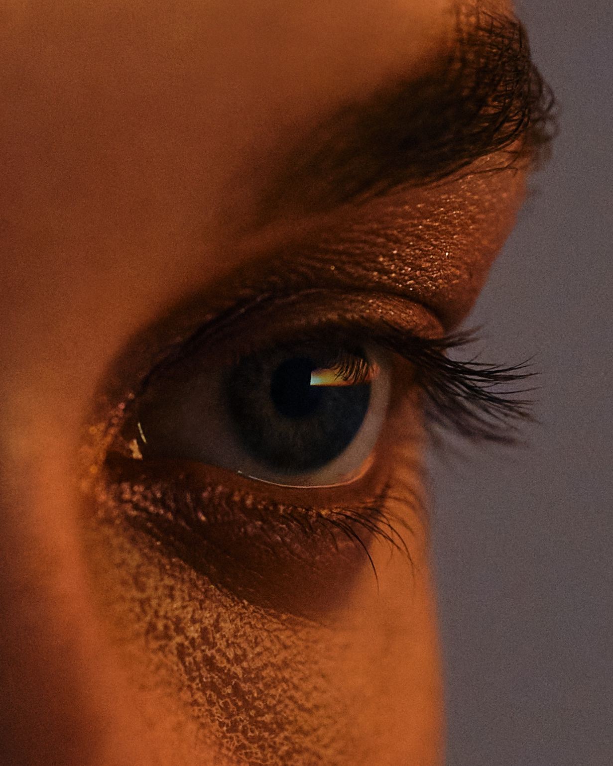 Extreme close-up of a human eye, lit warm orange from the left, with lashes and skin texture in sharp detail against a grey background.