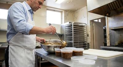 Cook ladling stew from a stainless bowl into a row of clear deli containers on a prep table