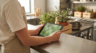 Cook in an apron holding a tablet at a stainless counter lined with potted herbs