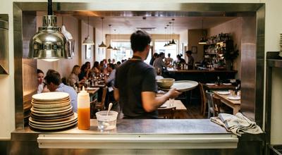 View through a kitchen pass into a busy dining room, with stacked plates and a squeeze bottle on the counter