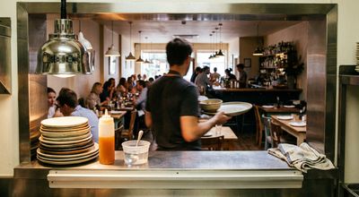View through a kitchen pass into a busy dining room, with stacked plates and a squeeze bottle on the counter
