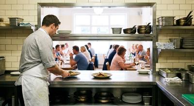 Cook setting plated dishes on a stainless pass shelf, a full dining room visible through the opening beyond