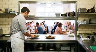 Cook setting plated dishes on a stainless pass shelf, a full dining room visible through the opening beyond