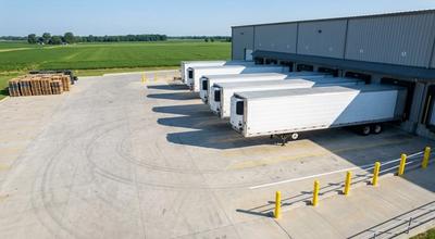 Refrigerated trailers backed into loading dock bays at a distribution warehouse, farm fields beyond