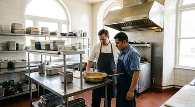 Two cooks looking over a pan of stew together at a stainless prep table in a bright tiled kitchen