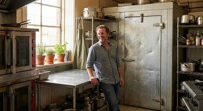 Man leaning on a prep table beside a weathered walk-in cooler door in an older commercial kitchen