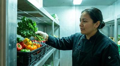 Cook in a black coat lifting kale from a produce crate on a shelf inside a walk-in cooler