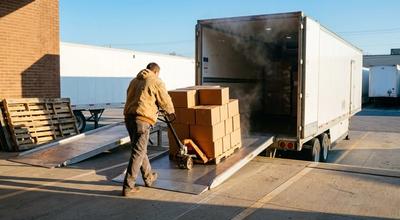 Worker pushing a pallet jack of boxed product up a ramp into a refrigerated trailer at a loading dock in low sun