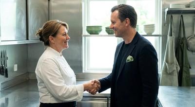 Chef in a white coat shaking hands with a man in a suit jacket in a stainless commercial kitchen