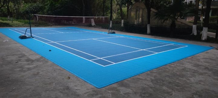 An outdoor badminton court laid in blue modular tiles, net up, marked with both singles and doubles lines