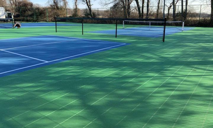 Two outdoor tennis courts surfaced in green and blue modular tiles, nets up, seen from the baseline