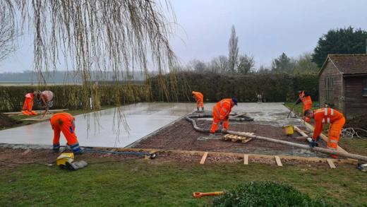 Two workers in hi-vis finishing a freshly poured concrete slab in a garden, ready to take a court.