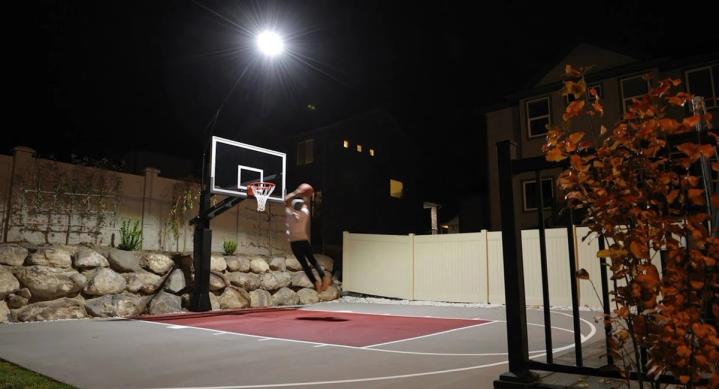 A floodlit home court at night, a player driving to the hoop under a single mast light beside a stone retaining wall.