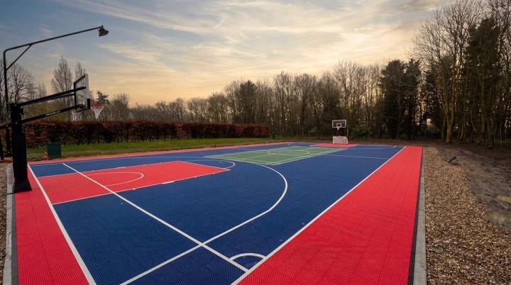 Egy teljes méretű, kék és piros multifunkciós sportpálya oldalnézete tollaslabda-betéttel és kosárlabda-gyűrűkkel naplementekor