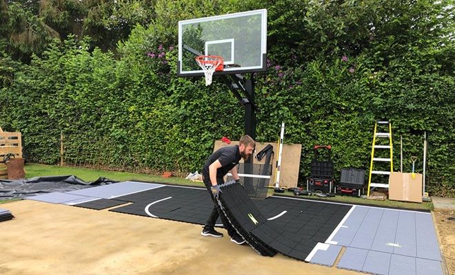 An installer laying black modular tiles across a prepared base in a garden, with the hoop already standing at the end of the court.