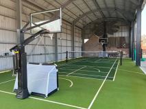 An indoor court inside a steel-framed building: green tiles marked for several sports, a net strung across the middle and a hoop at each end.