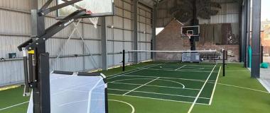 An indoor court inside a steel-framed building: green tiles marked for several sports, a net strung across the middle and a hoop at each end.