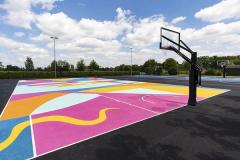 An outdoor school court surfaced in a bright pink, yellow and blue graphic pattern, with a black post-mounted hoop and floodlights behind.