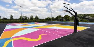An outdoor school court surfaced in a bright pink, yellow and blue graphic pattern, with a black post-mounted hoop and floodlights behind.