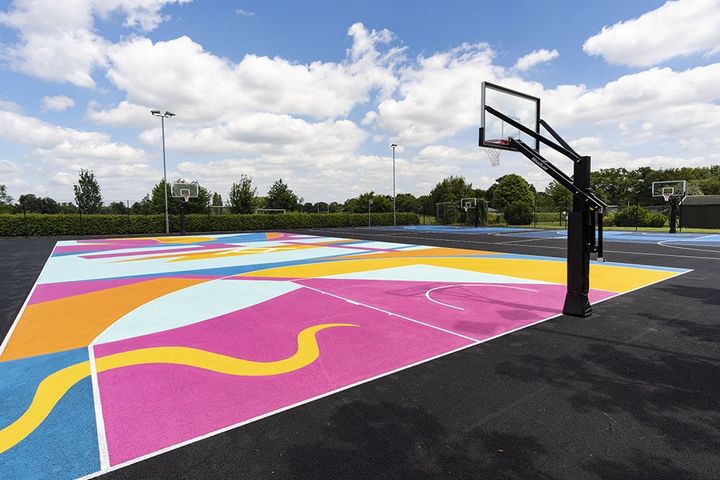 An outdoor school court surfaced in a bright pink, yellow and blue graphic pattern, with a black post-mounted hoop and floodlights behind.