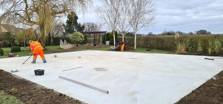 Two workers in hi-vis finishing a freshly poured concrete slab in a garden, ready to take a court.