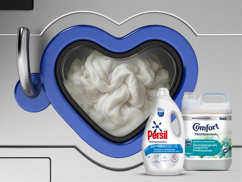 A large bottle of Persil Professional Non Bio laundry detergent stands in front of a washing machine with a blue door and white laundry, with another detergent bottle partially visible on the right.