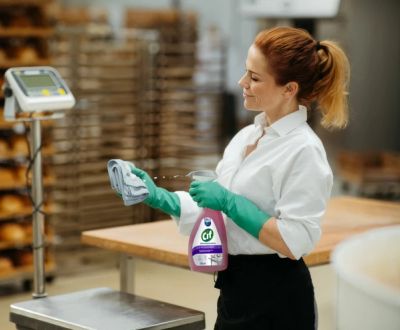 A smiling woman in green gloves sprays Cif cleaner onto a cloth in a professional kitchen or bakery.