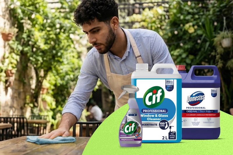 En man i förkläde torkar av ett bord utomhus, med Cif och Domestos Professional rengöringsprodukter i förgrunden.