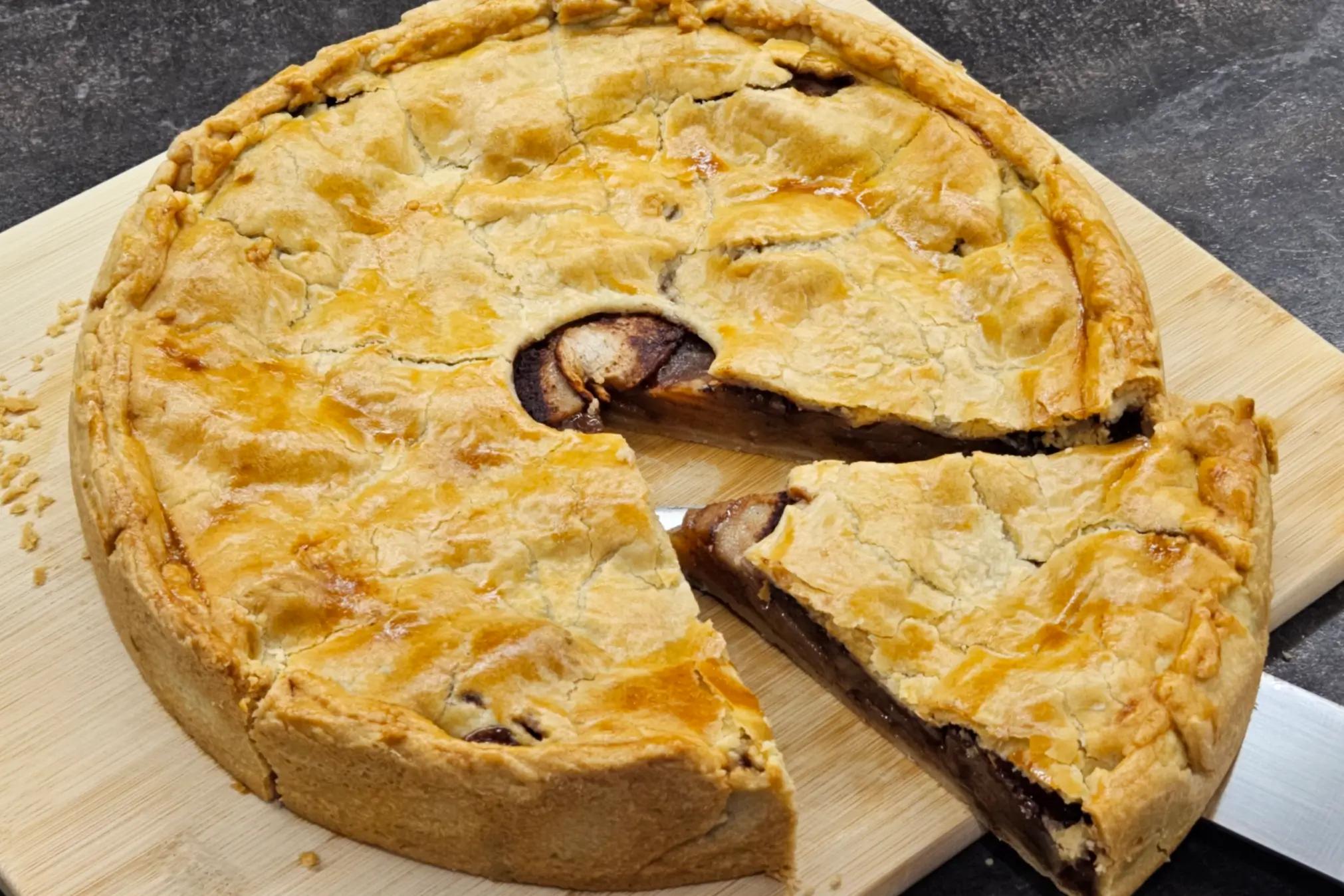 Круглий пиріг із золотисто-коричневою скоринкою. Один великий шматок відрізано й злегка відсунено, видно начинку з запечених яблук у карамельному соусі з корицею.