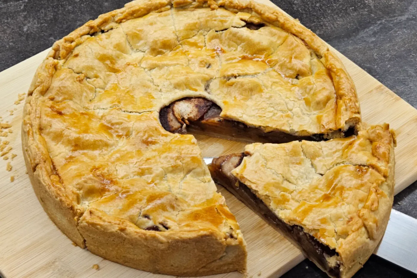 A round pie with a golden-brown crust. One large slice has been cut and slightly pulled aside, revealing a filling of baked apples in a caramel cinnamon sauce.