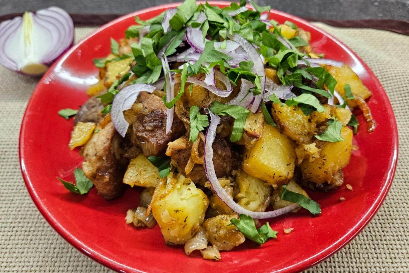 Roasted potatoes with meat and caramelized onions, topped with fresh herbs and slices of red onion, served on a red plate over a woven napkin.