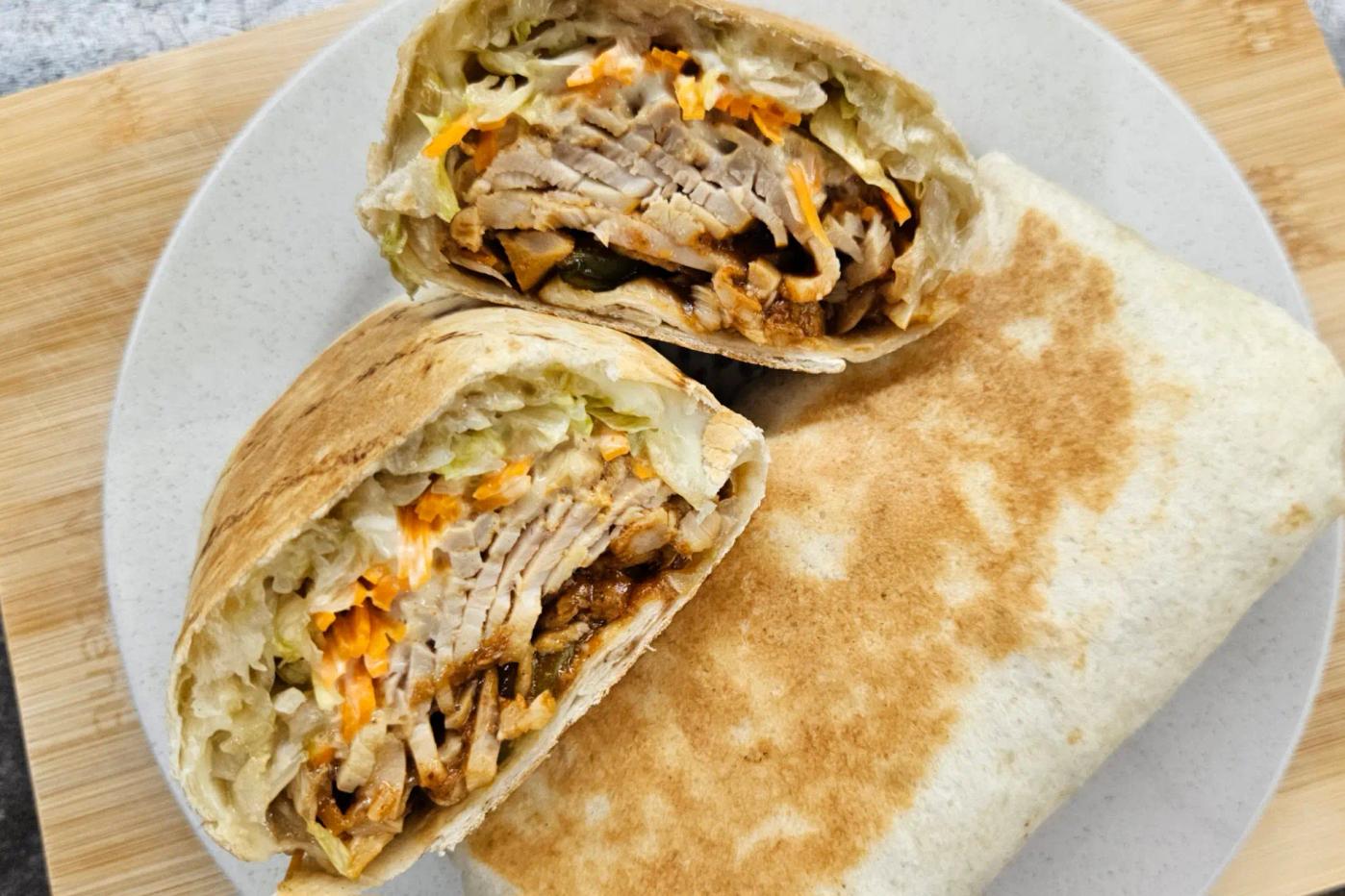 A warmed tortilla roll, cut in half, filled with juicy chicken, shredded lettuce, and carrot, served on a white plate set on a wooden board.
