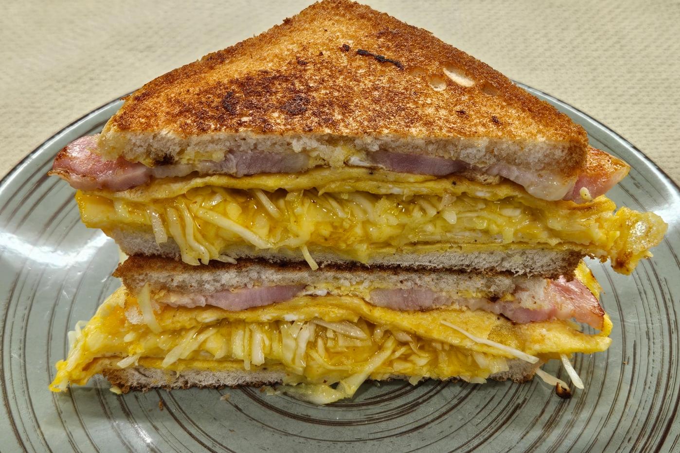 A sandwich with toasted bread, an omelet, ham, crunchy cabbage, and melted cheese, cut in half and stacked on a green plate.