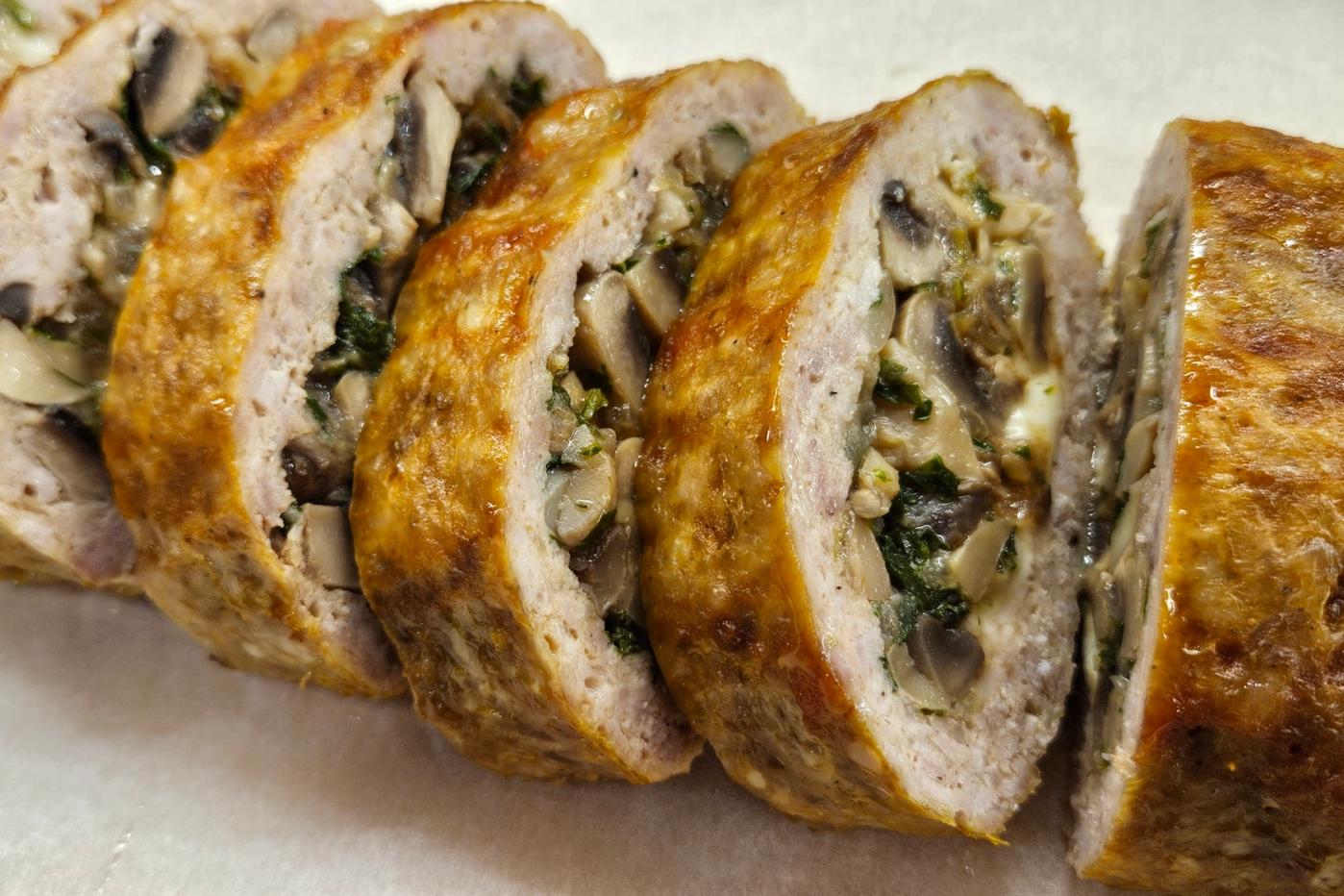 A stuffed meat roll with a golden crust, filled with mushrooms, herbs, and mozzarella, sliced and served on parchment paper.