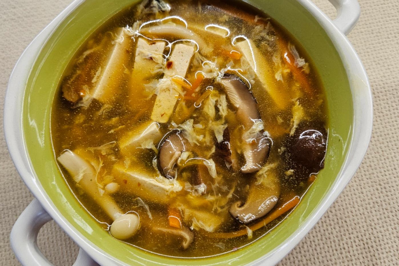 A deep bowl with two handles, filled with hot, dark broth soup with mushrooms, tofu, carrots, and egg.