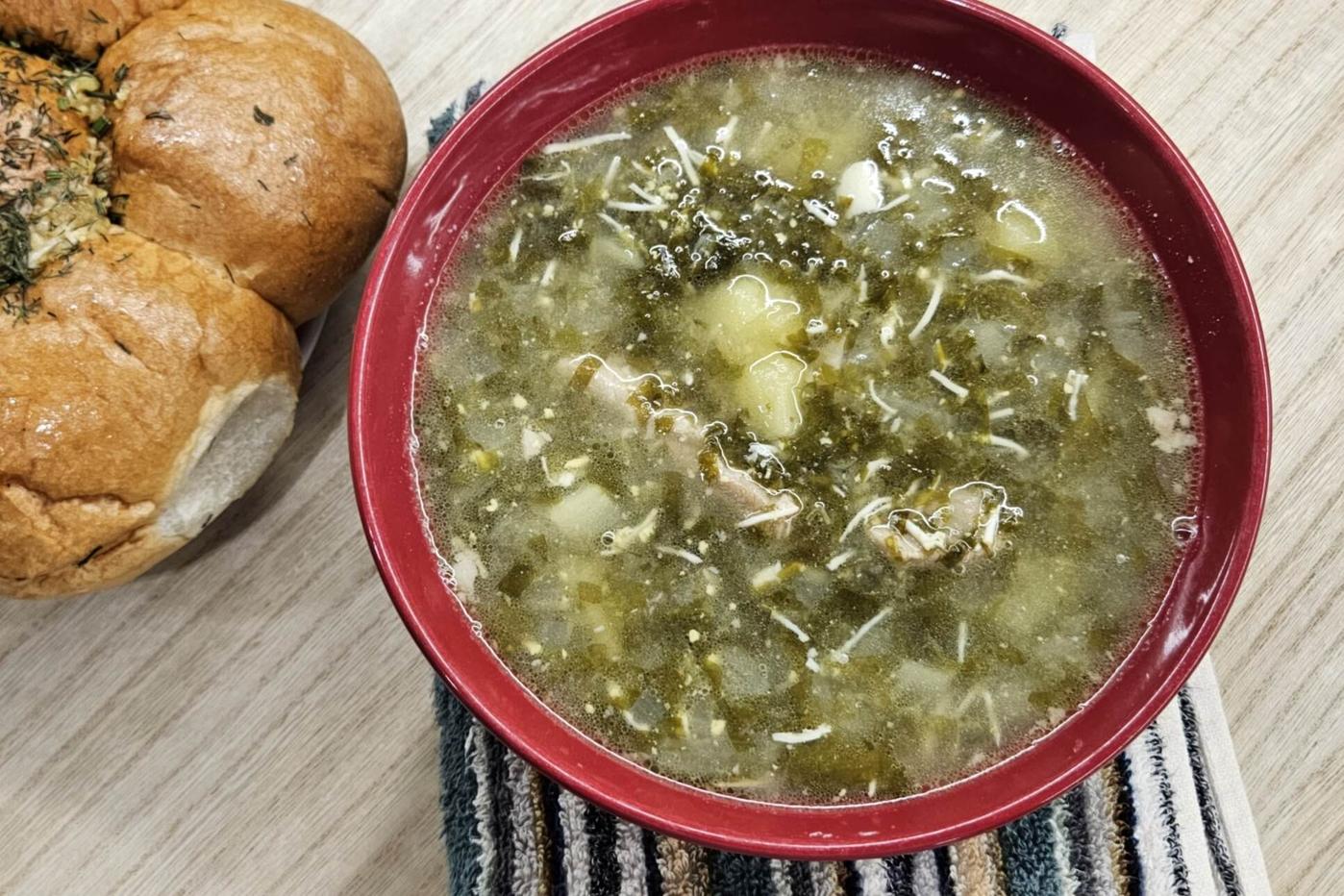 A red bowl filled with hot green borscht, showcasing tender potatoes and fresh greens, served alongside soft, fluffy garlic buns sprinkled with herbs.