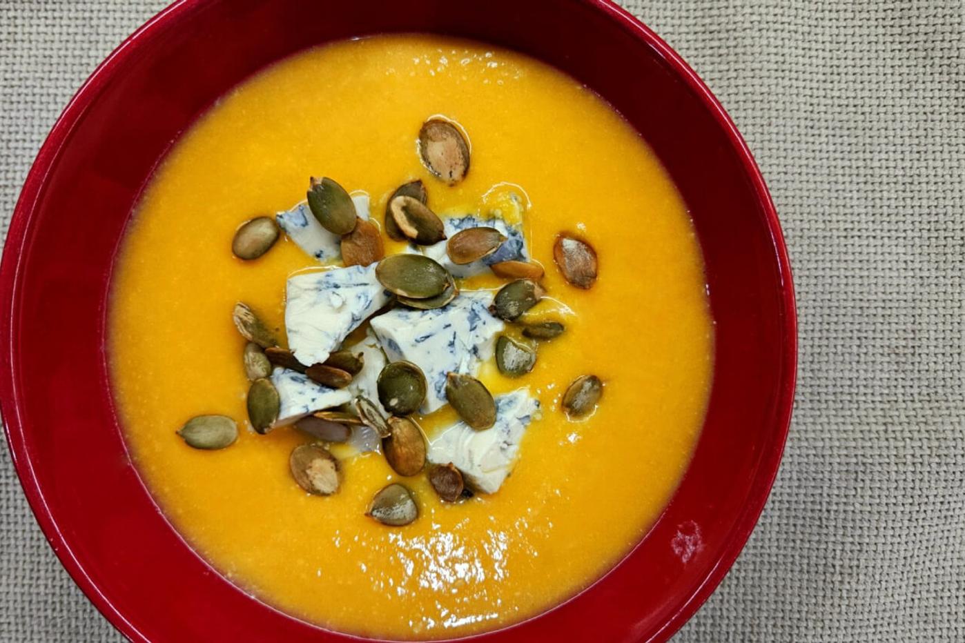 Creamy pumpkin soup served in a red bowl, topped with Dorblu cheese and sunflower seeds.