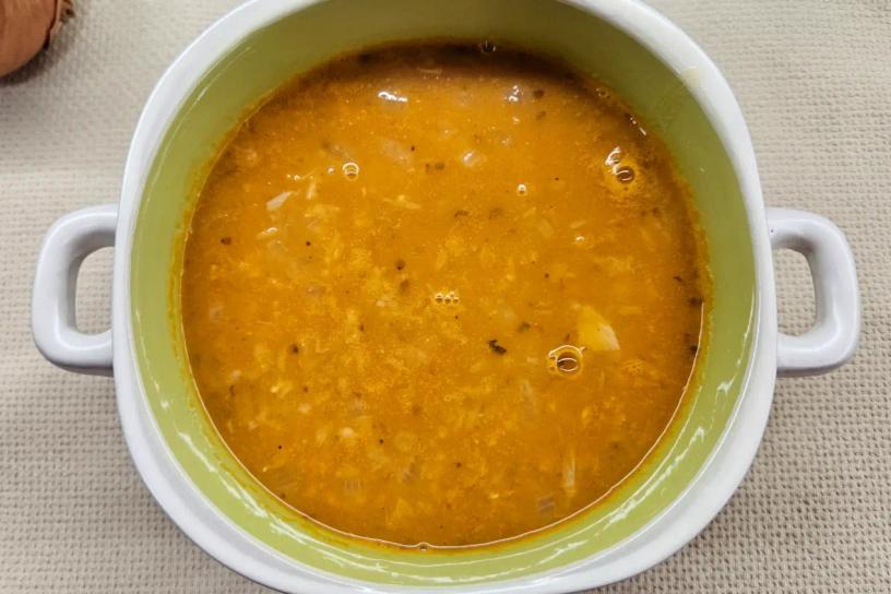 A deep white bowl with two handles filled with thick orange-brown soup, speckled with small vegetable pieces and spices, placed on beige fabric.