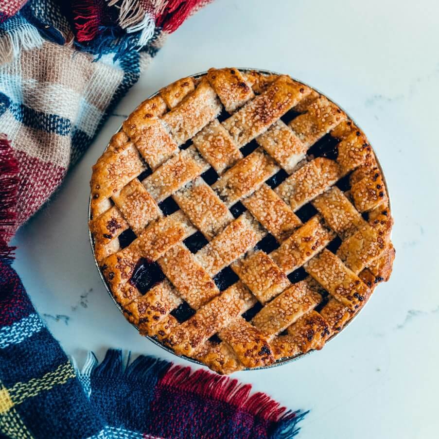 Золотисто-коричневий ґратчастий пиріг, посипаний цукром, начинкою з темних фруктів, на білій поверхні, поряд із кольоровою картатою тканиною.